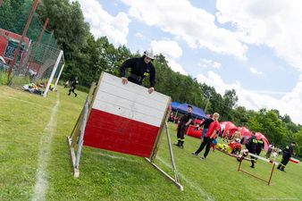 strażak w czarnym mundurze i białym hełmie wspina się na wysoką biało-czerwoną przeszkodę 