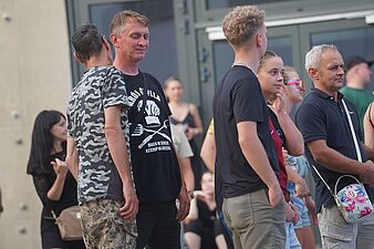 Mężczyzna w czarnej koszulce z nadrukiem i inni uczestnicy obserwujący imprezę plenerową