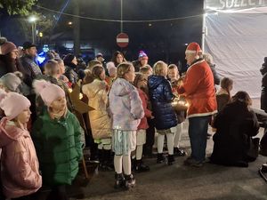 Święty Mikołaj rozmawia z dziećmi na zewnątrz, podczas gdy grupa ludzi obserwuje wydarzenie.