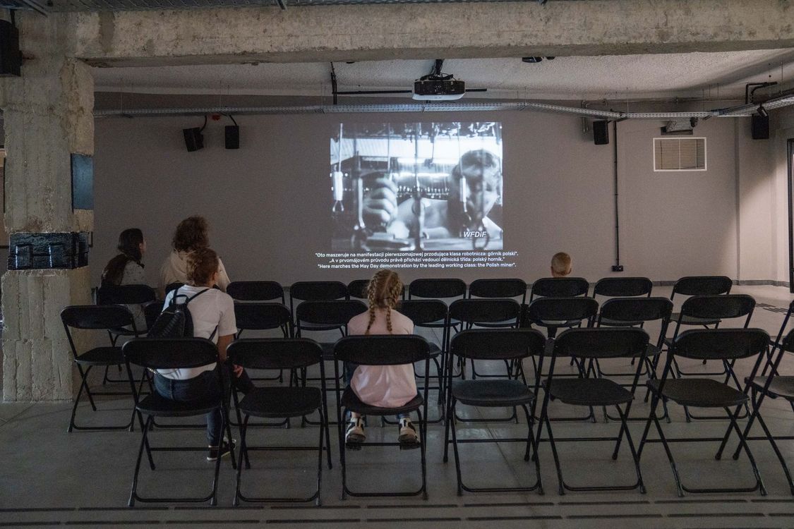 Niewielka sala kinowa na wystawie, w której zwiedzający siedzą na krzesłach i oglądają archiwalny, czarno-biały film dokumentalny wyświetlany na ścianie