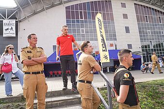 Grupa osób, w tym strażacy oraz mężczyźni w koszulkach reprezentacyjnych, stoi na schodach budynku sportowego i obserwuje pokazy ratownictwa