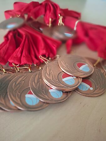 Zbliżenie na pęk miedzianych medali sportowych z czerwonymi wstążkami. Na medalach widnieje naklejka z motywem piłki w mikołajkowej czapce.