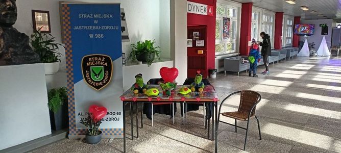 Stoisko Straży Miejskiej w Jastrzębiu-Zdroju na WOŚP, umiejscowione w holu budynku. Na stole widać maskotki, żółte czapki i serca. Obok stoi baner, wiklinowe krzesło oraz popiersie. W oddali widać przechodniów i monitory