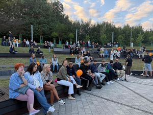17. Tłum ludzi siedzi na ławkach patrząc w różnych kierunkach. Niektórzy trzymają pomarańczowe balony. 