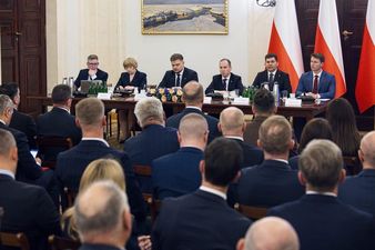 6osób siedzi przy stole prezydialnym w ciemnych ubraniach za nimi jasna ściana z obrazem po prawej stronie flagi Polski