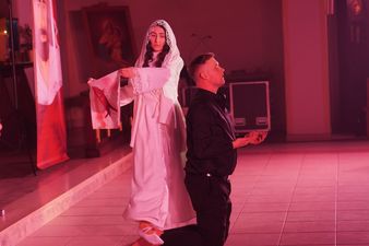 Klęczący mężczyzna utożsamiający postać Księdza Machy, za Nim stojąca kobieta ubrana na biało trzyma w rękach chustę z wizerunkiem Chrystusa