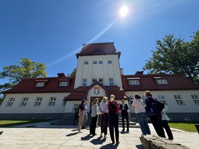 Grupa ludzi rozmawia na placu przed głównym wejściem do zabytkowego gmachu w słoneczny dzień