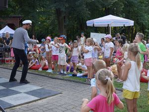 Animator w pasiastej koszulce i marynarskiej czapce prowadzi zabawy taneczne dla grupy dzieci zgromadzonych w parku