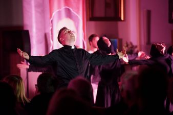 Aktor ubrany w sutannę utożsamiający postać księdza Machy z rozłożonymi ramionami spogląda w górę, wokół inne postaci ubrane na czarno, z przodu widownia