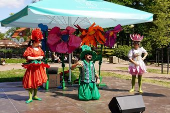 Kobiety i mężczyzna bajkowych strojach bawią widzów, na nimi kolorowe sztuczne kwiaty oraz parasol z napisem Jastrzębie-Zdrój