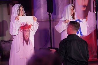 Dwie aktorki ubrane na biało trzymają rysunki twarzy Jezusa Chrystusa, przed nimi klęczący mężczyzna utożsamiający księdza Jana Machy