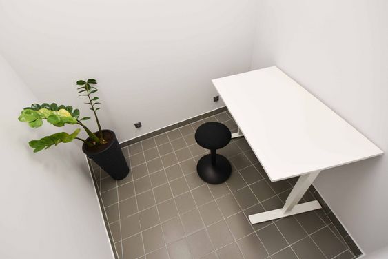 Małe, minimalistyczne biuro: białe biurko, czarny hoker i wysoka roślina doniczkowa (zamiokulkas) w czarnej donicy. Jasne ściany i szara podłoga z płytek. Nowoczesny, surowy wystrój