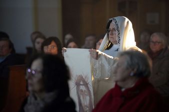 Kobieta ubrana na biało trzyma w rękach chustę z wizerunkiem Chrystusa, wokół zgromadzenie widzowie