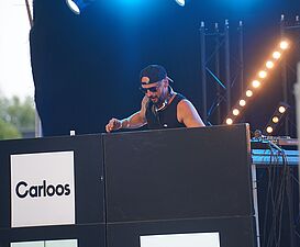 DJ w czarnej koszulce, okularach przeciwsłonecznych i czapce tyłem do przodu grający na konsoli z logo „Carloos”