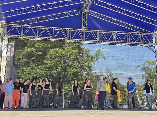 Grupa młodzieży w luźnych strojach zebrana na scenie pod dużym, niebieskim dachem amfiteatru