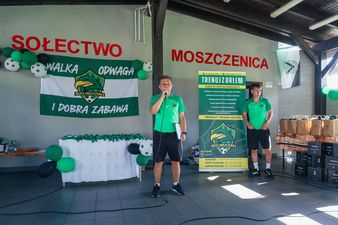 Oficjalna część festynu, podczas której trener w zielonej koszulce przemawia przez mikrofon, stojąc obok roll-upa klubu UKS Orzeł Moszczenica