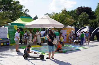 Kolorowa mata z tematyką ekologiczną, przed nią ustawiona hulajnoga, na drugim planie kolrowe namioty z grupką osób