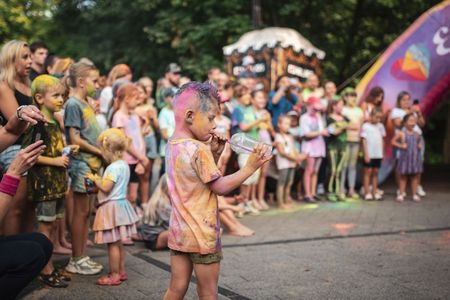 Chłopiec umazany kolorowym proszkiem holi pije napój przez słomkę, stojąc przed tłumem innych dzieci