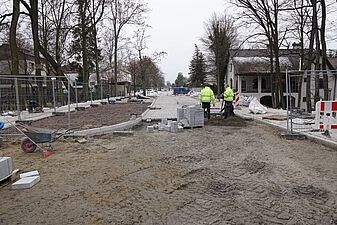 To szeroki kadr placu budowy przedstawionego na zdjęciu 5. Robotnicy układają kostkę, a pas ziemi po lewej stronie drogi został przygotowany pod zieleń lub chodnik. W głębi widać zarysy domów jednorodzinnych, drzewa i stojący, niebieski pojazd dostawczy
