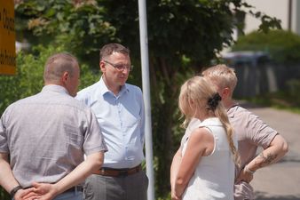 Ta sama grupa rozmawia na chodniku przy skrzyżowaniu dróg. W tle widać zaparkowane samochody i rząd wysokich drzew