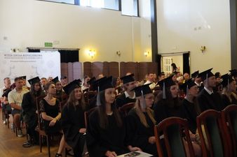 Studenci w czarnych togach siedzą na widowni. Po lewej stronie widoczna ścianka promocyjna uczelni z hasłem „Wartość Siła Bezpieczeństwo”