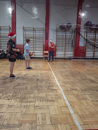 Dwoje uczniów stoi na parkiecie sali gimnastycznej podczas wydarzenia sportowego. W głębi sali, pod drabinkami, stoi mężczyzna w czerwonej koszulce i mikołajkowej czapce