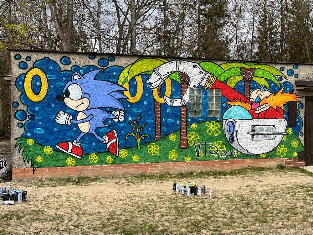 Kolorowy mural na ścianie przedstawiający postać Jeża Sonica biegnącego obok złocistych pierścieni i doktora Eggmana w swoim pojeździe