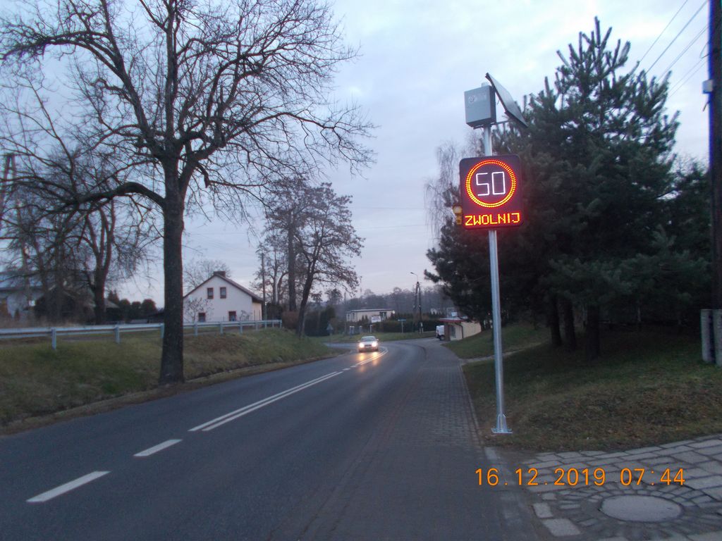 Elektroniczna tablica do mierzenia prędkości przy ulicy, w oddali samochód, przystanek i domy