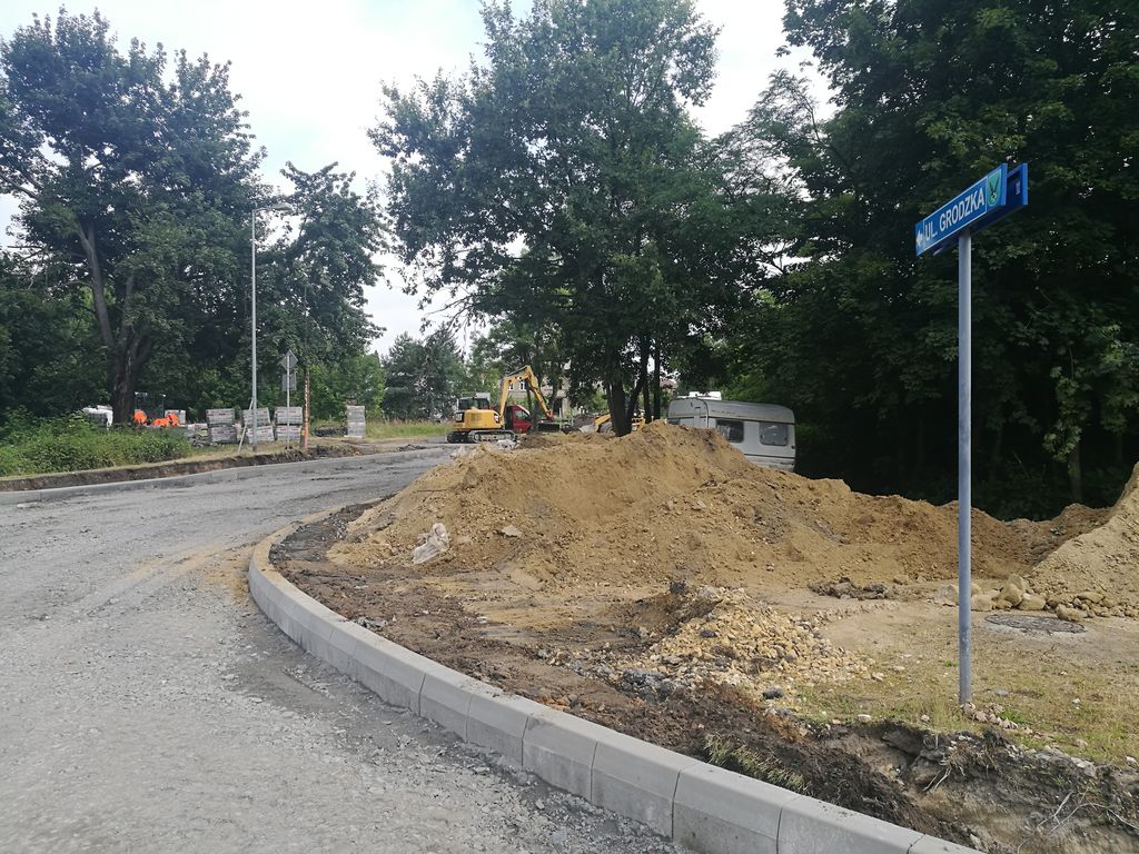 Plac budowy drogi przy ul. Grodzkiej. Na pierwszym planie hałda ziemi i nowe krawężniki wyznaczające łuk jezdni. W tle żółta koparka, tabliczka z nazwą ulicy i drzewa. Droga jest w trakcie korytowania i przygotowywania podbudowy