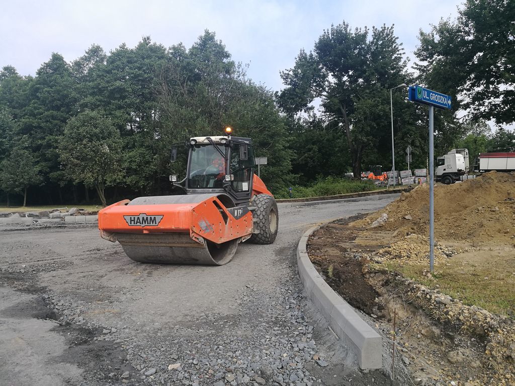 Pomarańczowy walec drogowy marki Hamm pracuje na placu budowy przy ul. Grodzkiej. Maszyna zagęszcza kruszywo obok nowo osadzonych krawężników. W tle widoczna tabliczka z nazwą ulicy, ciężarówka i ściana lasu