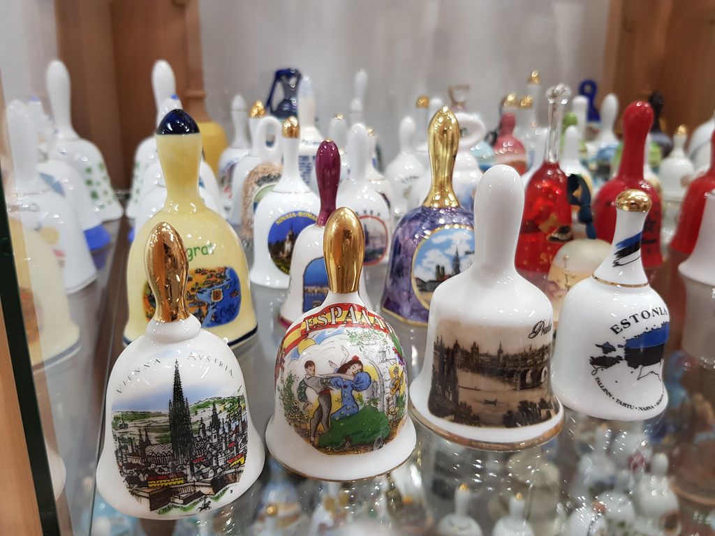 olekcja ceramicznych dzwonków pamiątkowych ustawionych na szklanej półce. Na dzwonkach widnieją kolorowe grafiki i napisy z różnych krajów i miast, m.in. Wiedeń, Hiszpania, Estonia i Praga. Niektóre dzwonki mają zdobione, złote uchwyty.