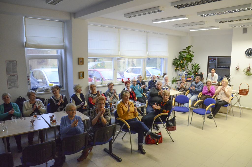 wnętrz sali gdzie obraduje klub seniora. Członkowie siedzą przy stolikach i klaszczą