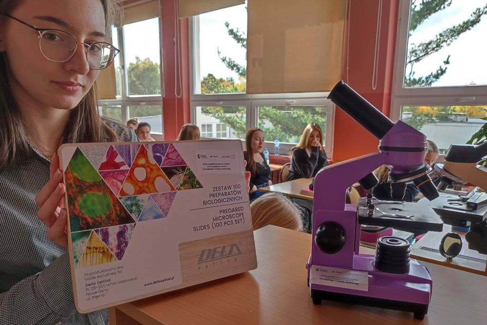 Uczennica prezentuje mikroskop i zestaw preparatów.