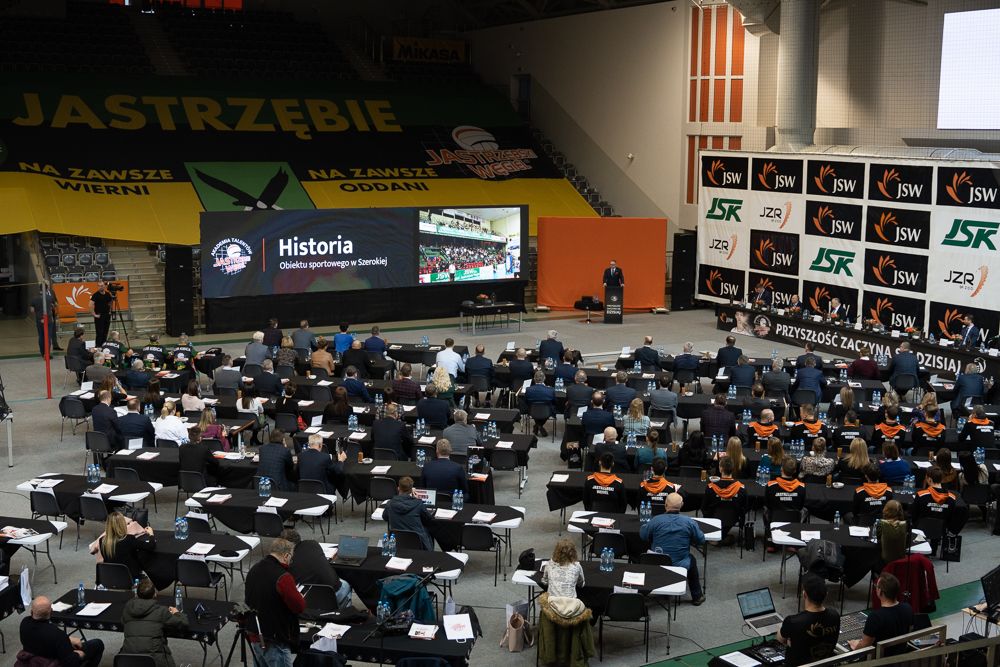 Widok z trybun na płytę hali - widać mnóstwo stolików, stół konferencyjny. Przemawia Adam Gorol.