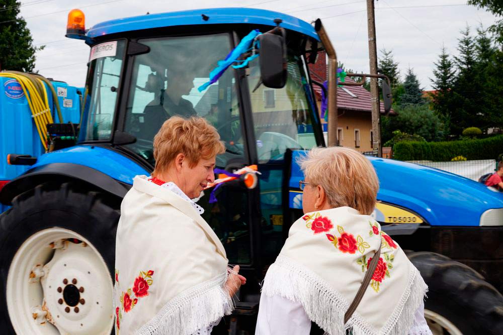 Zdjęcie przedstawia kobiety w strojach regionalnych na tle ciągnika rolniczego.