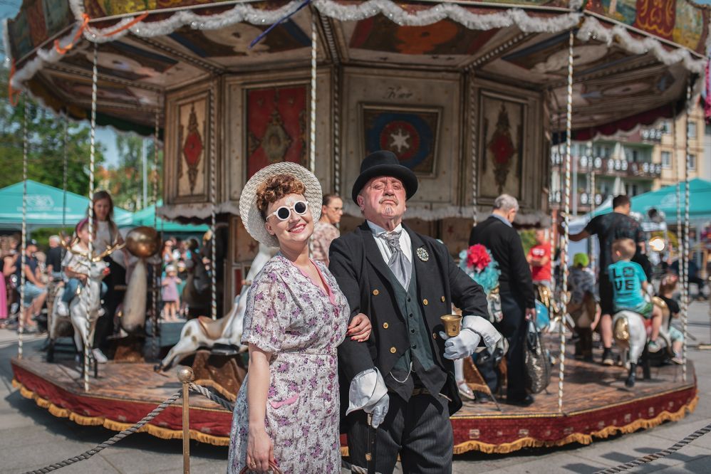 Zdjęcie przedstawia parkę ubraną w strój z epoki na tle karuzeli.