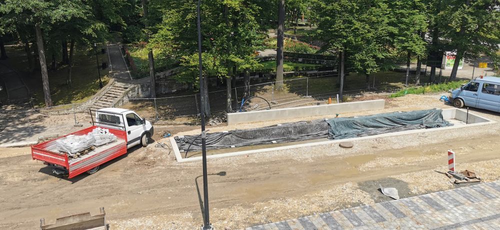 Widok z góry na plac budowy w parku. Obok betonowej wnęki osłoniętej folią stoi biała ciężarówka z materiałami. W tle gęste korony drzew, alejki i ogrodzenie zabezpieczające teren prac