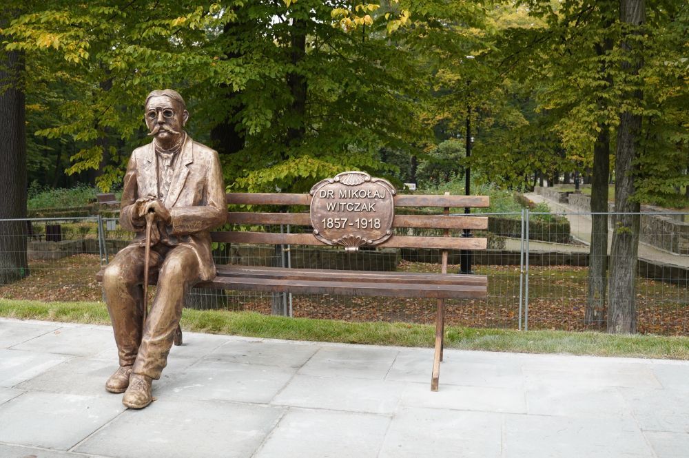 Zdjęcie przedstawia ławeczkę z rzeźbą Mikołaja Witczaka znajdującą się na deptaku w Zdroju.