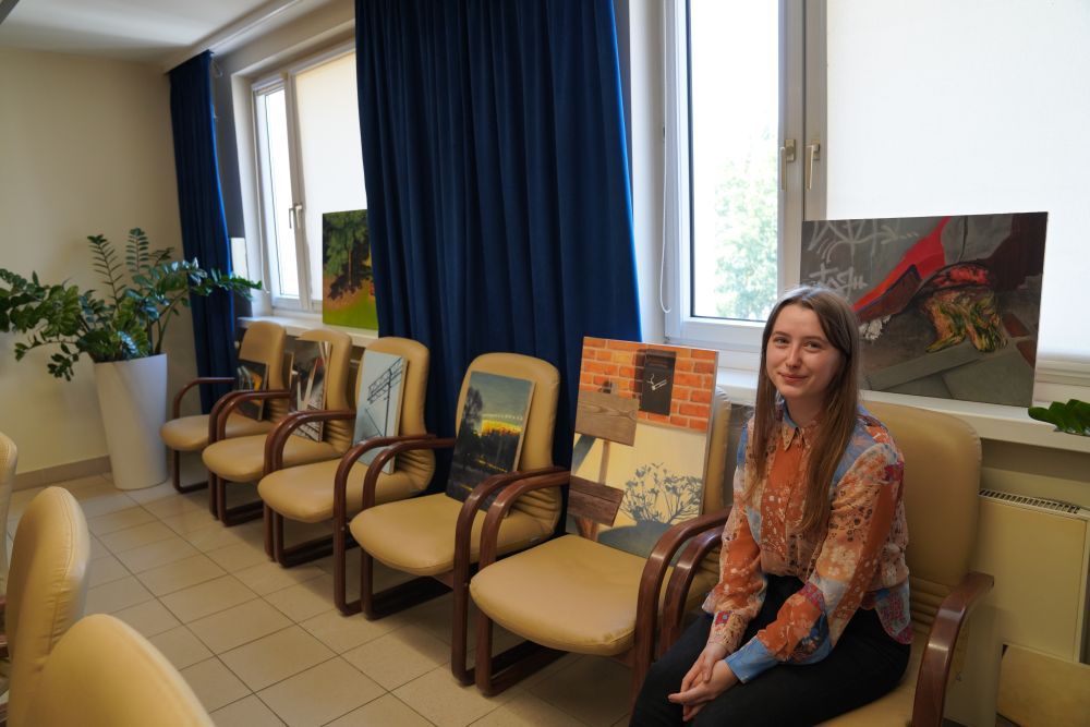 Zdjęcie przedstawia artystkę siedzacą obok swoich obrazów poukładanych na krzesłach. 