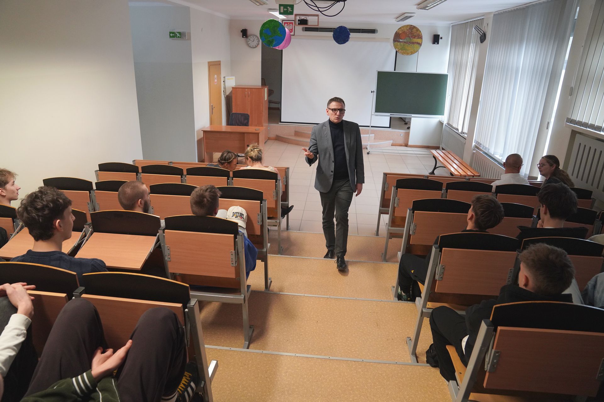 Uczniowe siedzący na auli, ujęcie z tyłu, do nich przemawia prezydent w tle biały ekran, tablica, zegar