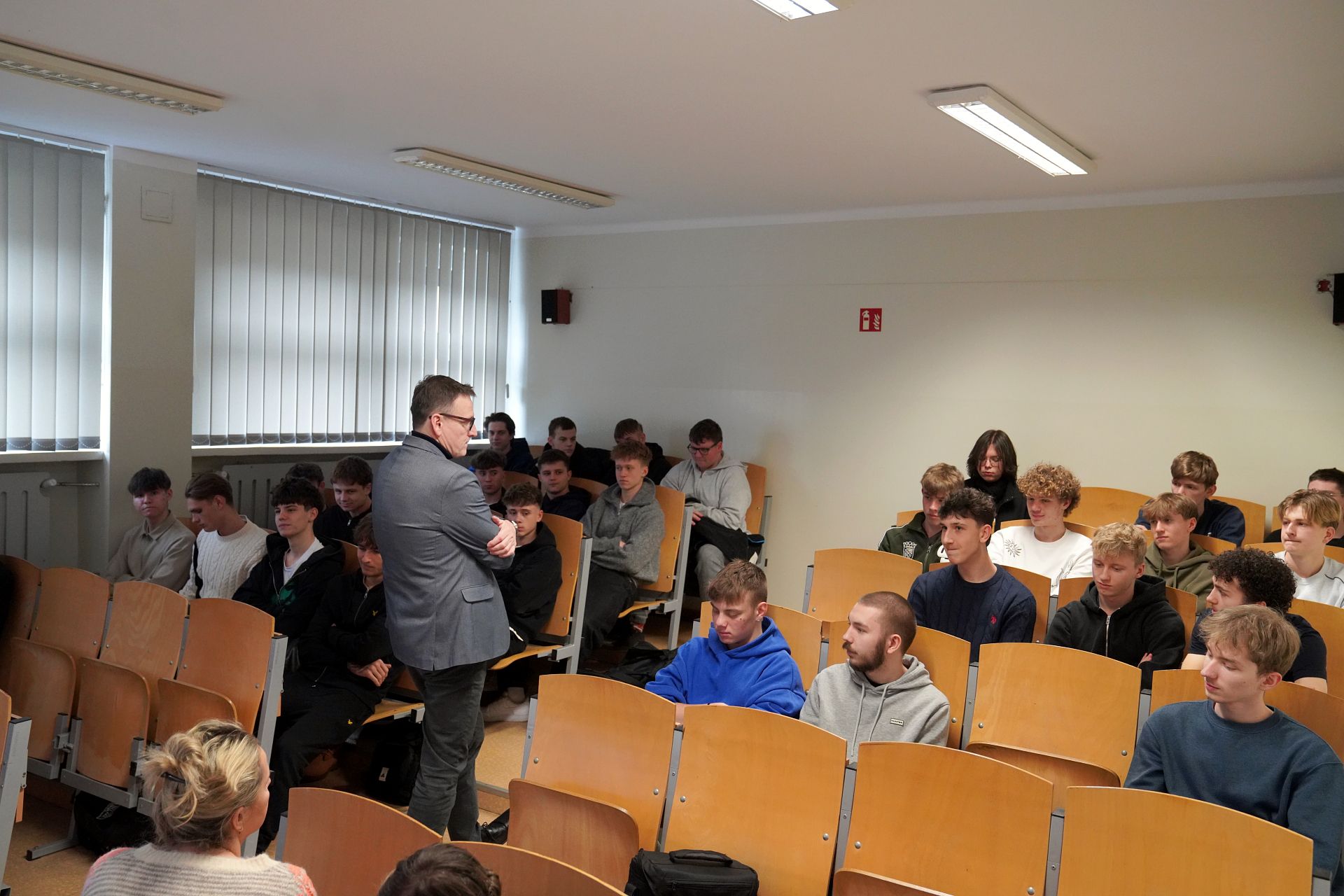 Uczniowe siedzący na auli, ujęcie z boku, do nich przemawia prezydent w tle ściana i okna zasłonięte roletami