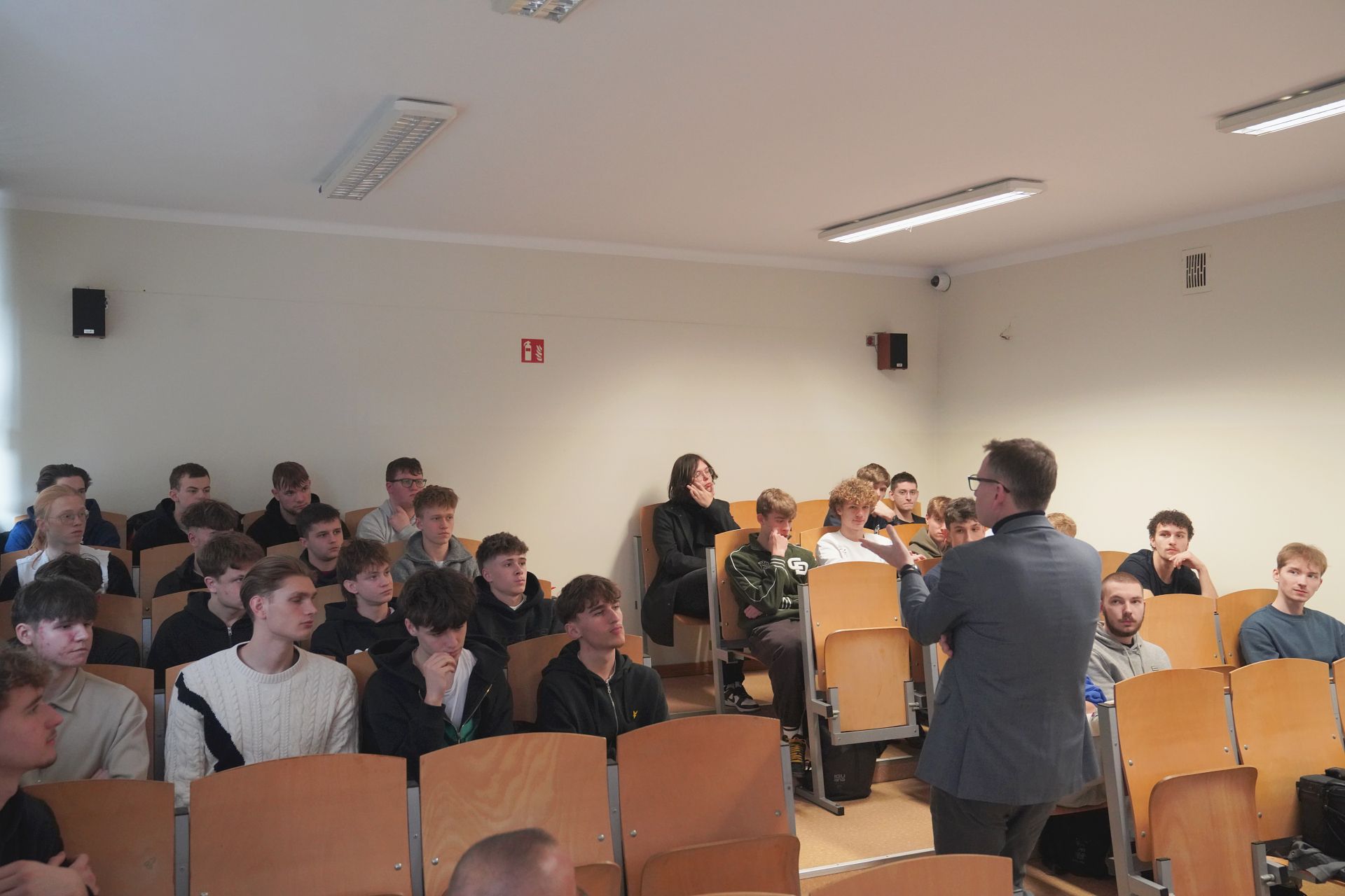 Uczniowe siedzący na auli, ujęcie z boku, do nich przemawia prezydent w tle ściana