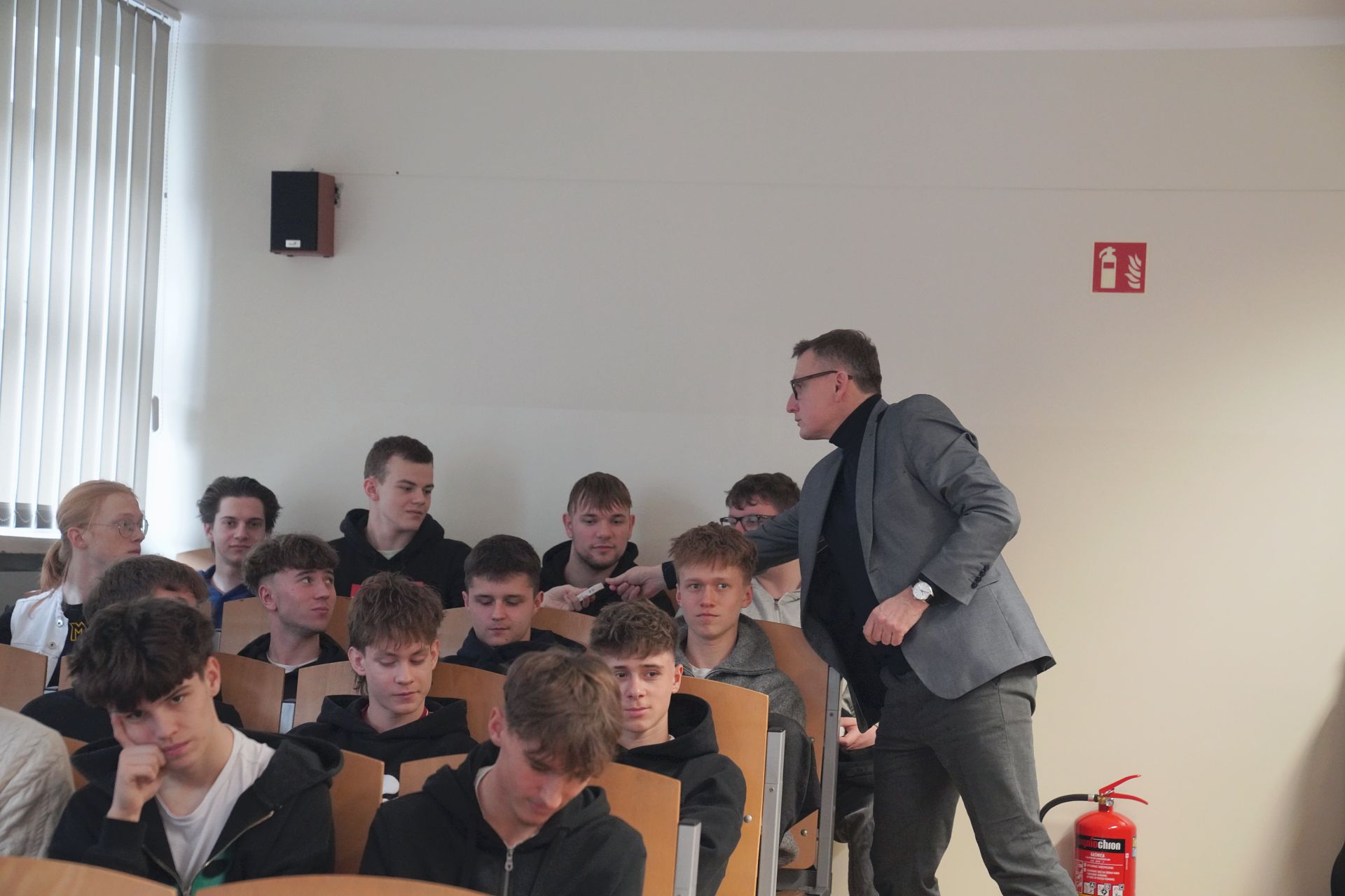 Uczniowe siedzący na auli, ujęcie z boku, prezydent jednemu uczniowi wręcza upominek w tle ściana i gaśnica na ziemi