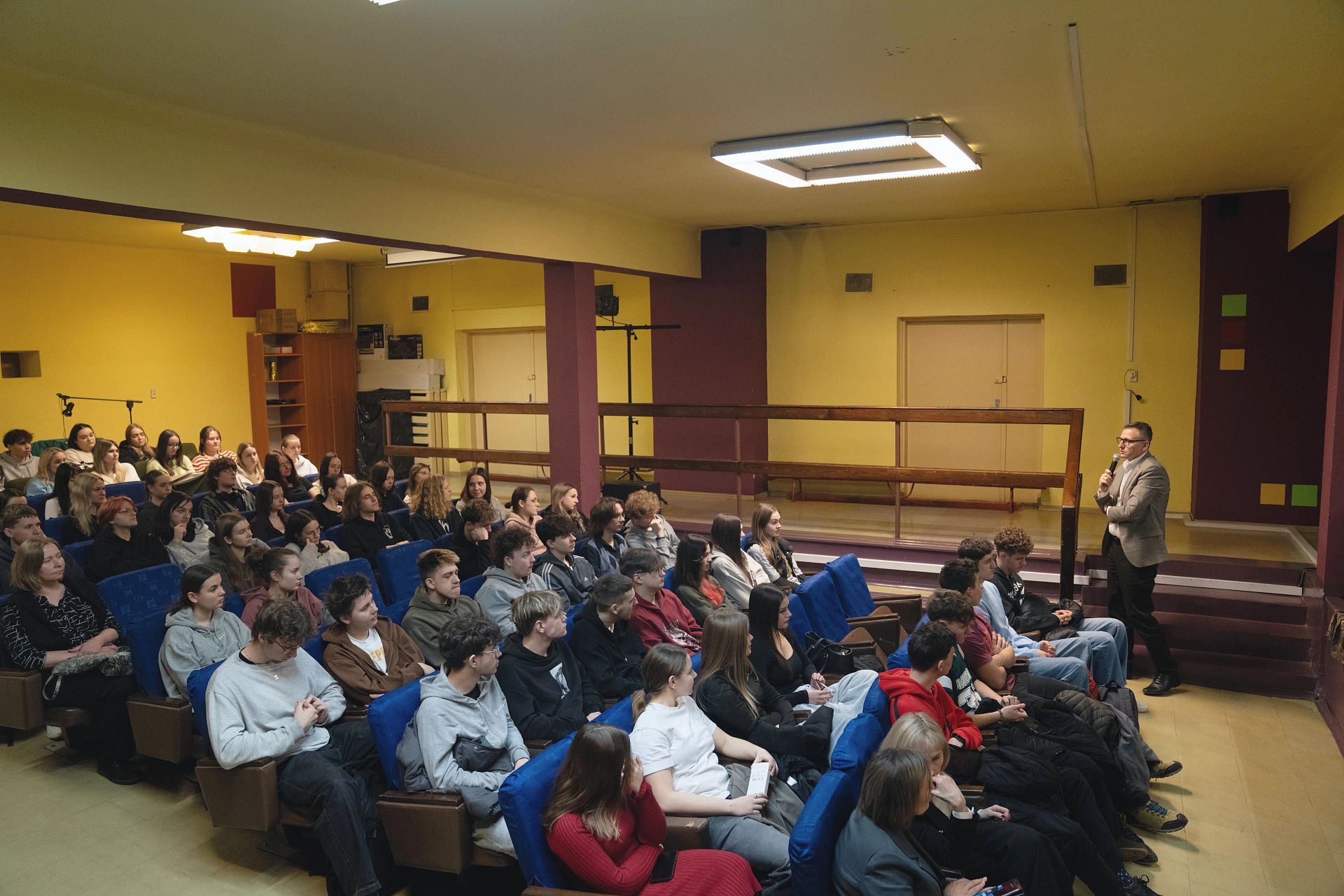 Uczniowe siedzący na auli, ujęcie z boku, do nich przemawia prezydent w tle drzwi i balustrada