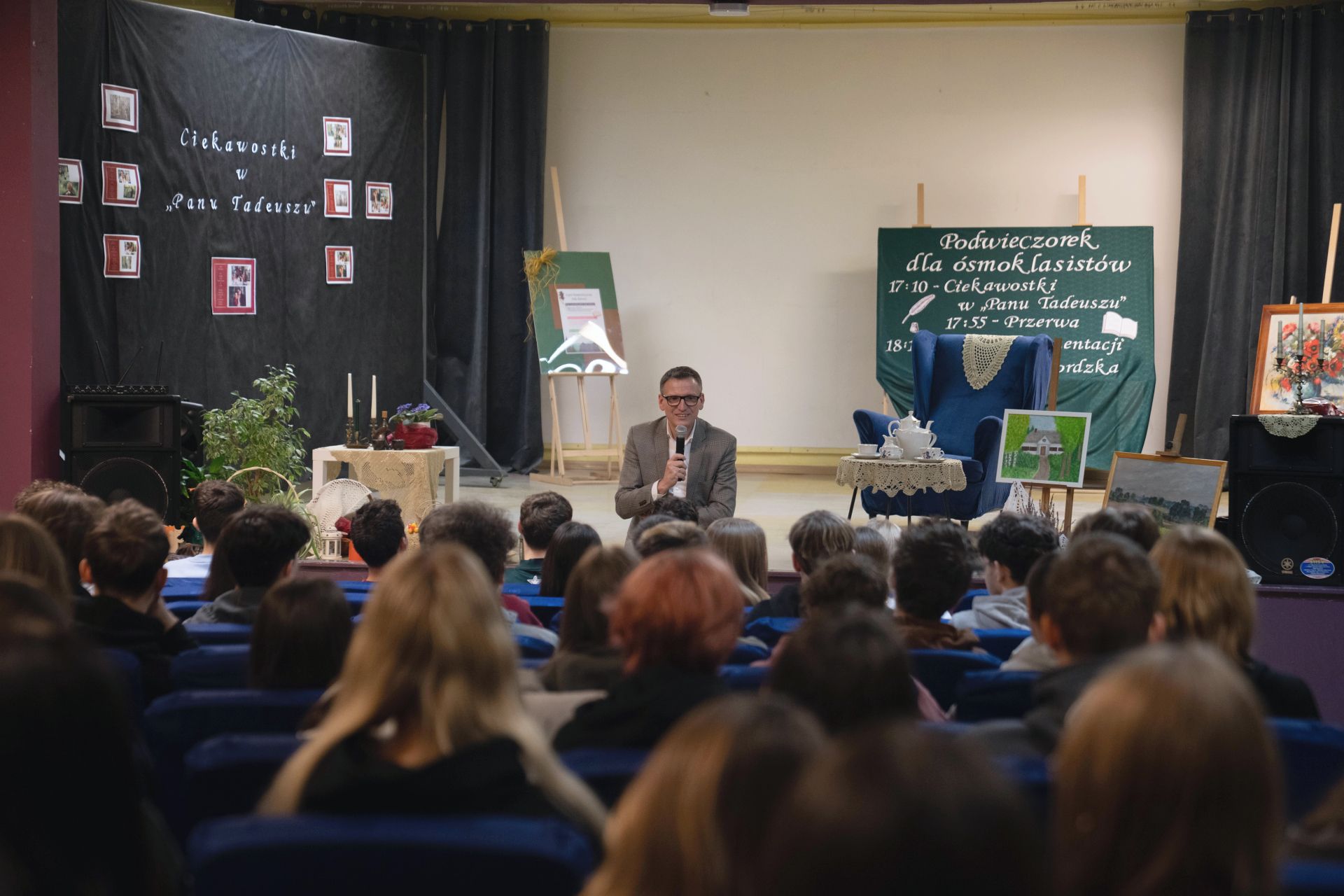 Uczniowe siedzący na auli, ujęcie z tyłu, do nich przemawia prezydent w tle scena a na niej fotel, obrazy, stolik ze świecami