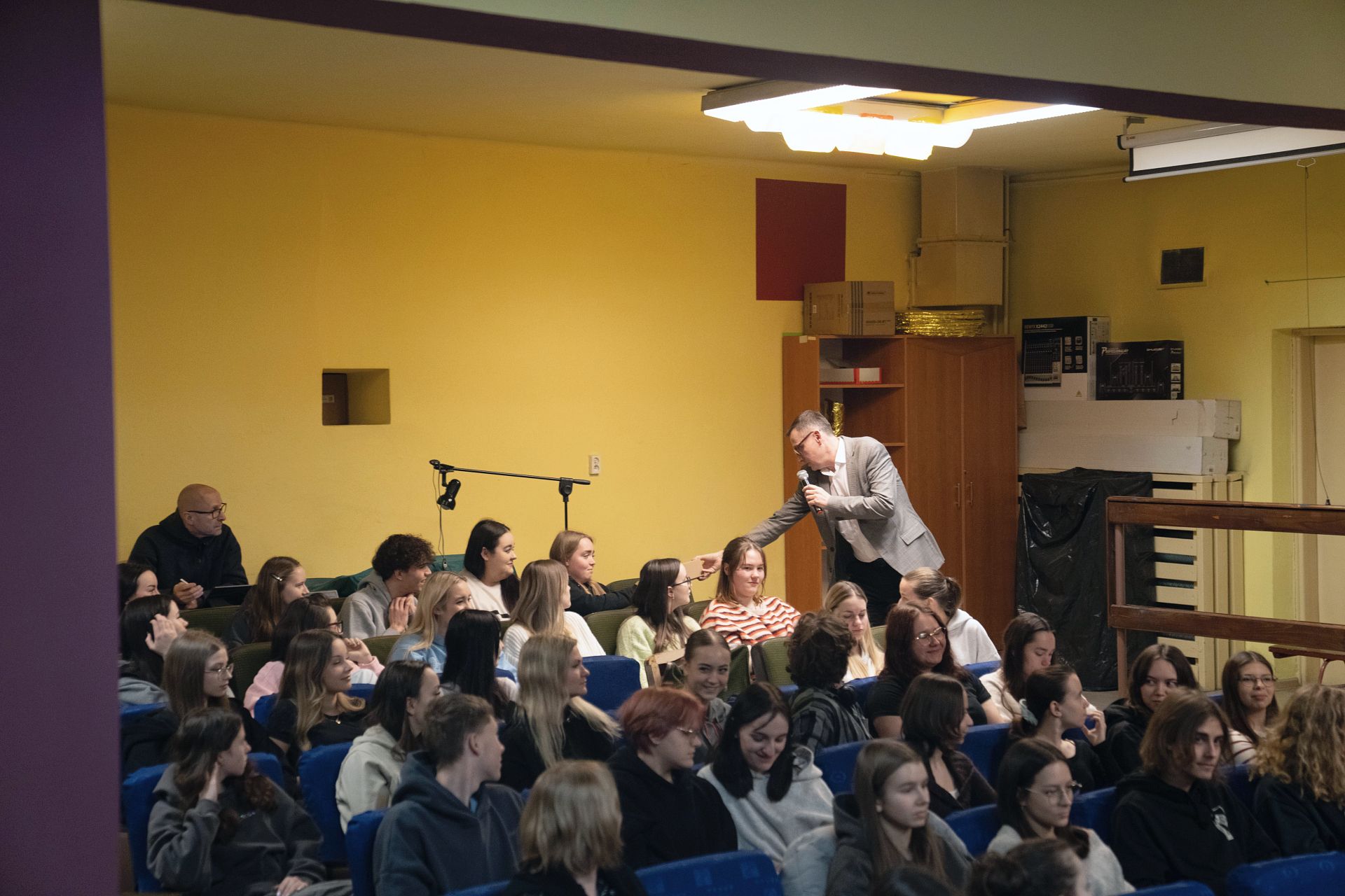 Uczniowe siedzący na auli, ujęcie z boku, prezydent jednej z uczennic wręcza upominek, w tle balustrada, szafa