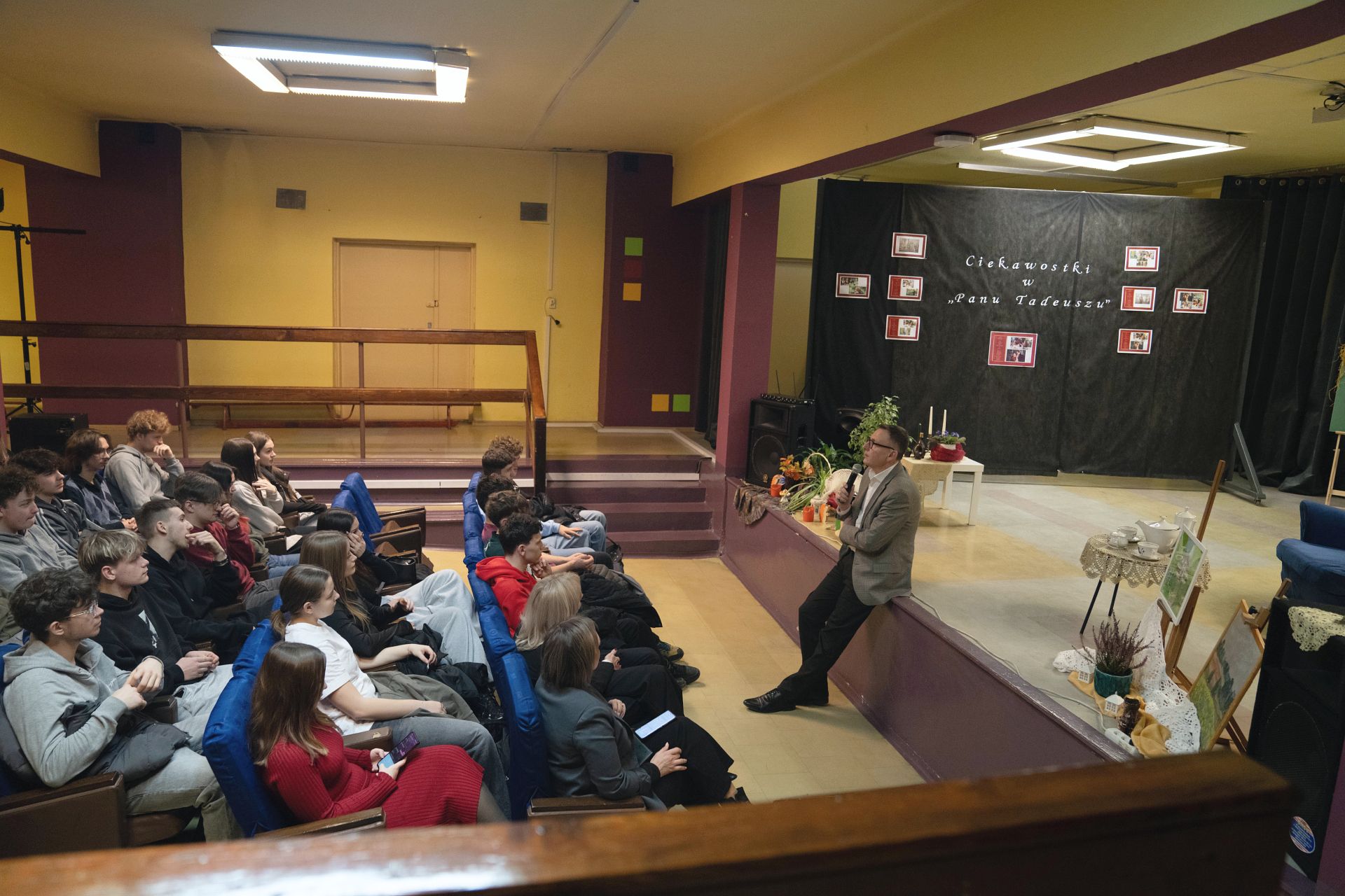 Uczniowe siedzący na auli, ujęcie z boku, do nich przemawia prezydent w tle drzwi i balustrada i fragment sceny