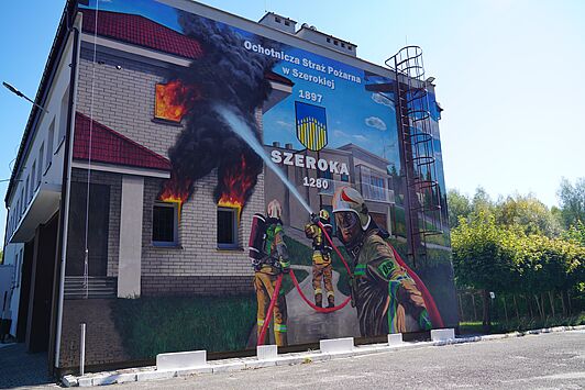 Mural na elewacji przedstawia strażaków gasiących pożar płonącego budynku. U góry widnieje napis „Ochotnicza Straż Pożarna w Szerokiej 1897”, herb oraz nazwa miejscowości „SZEROKA 1280”
