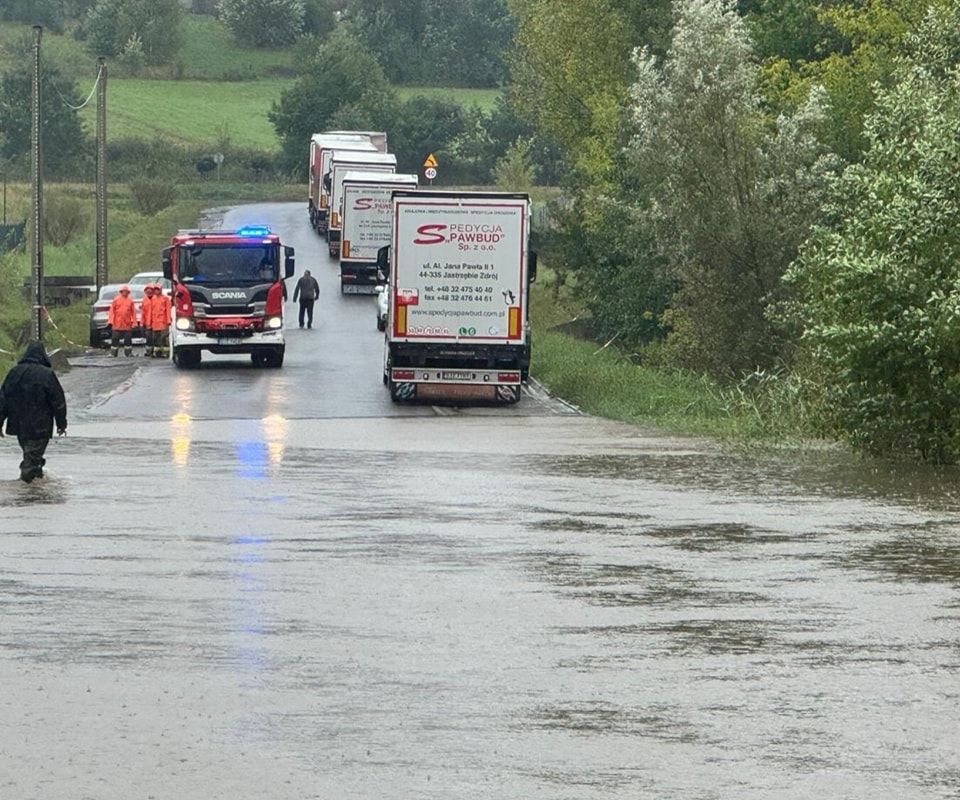 Zdjęcie przedstawia zalaną drogę, ciężarówki oraz ludzi.
